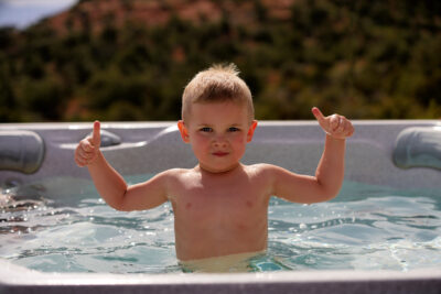 A young blonde boy is giving the thumbs up while in a hot tub outdoors.